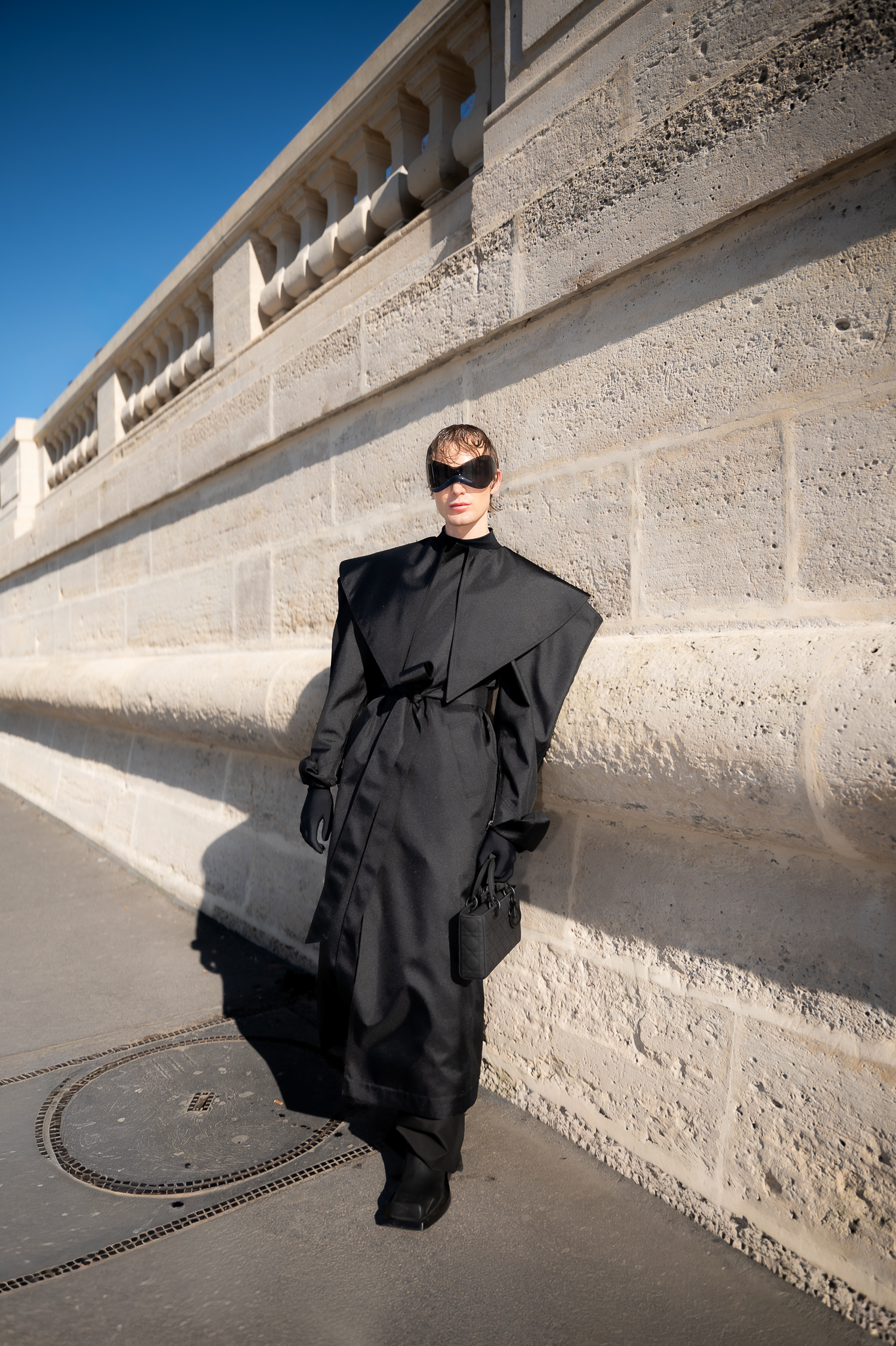 Client posing in a black fashion outfit by a stone wall during Paris Fashion Week