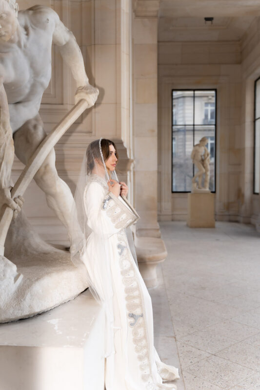 Woman wearing a veil and caftan posing near a sculpture during an editorial shoot in Paris